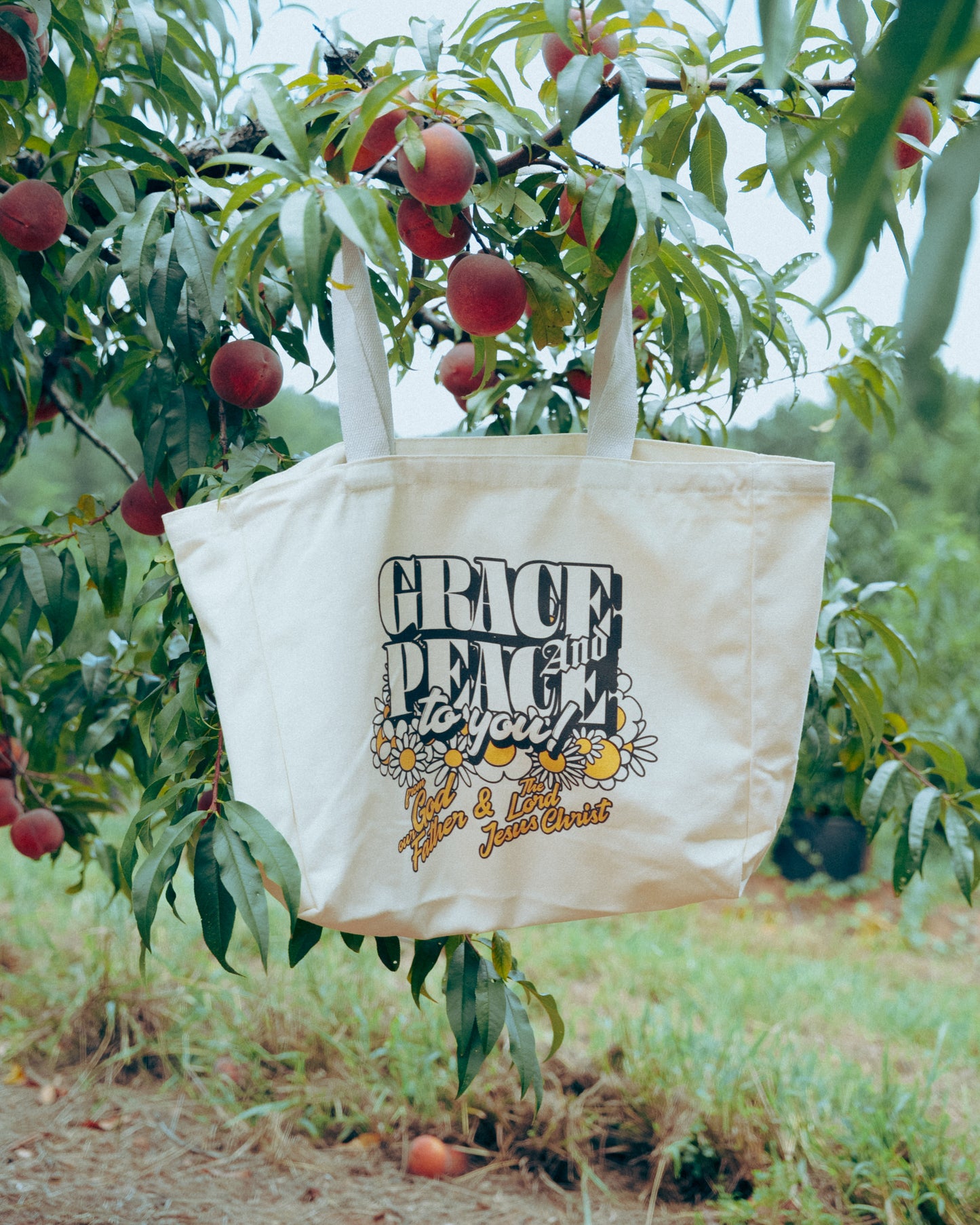 cream Tote bag with 'Grace, Peace, and Joy' text hanging from a peach tree