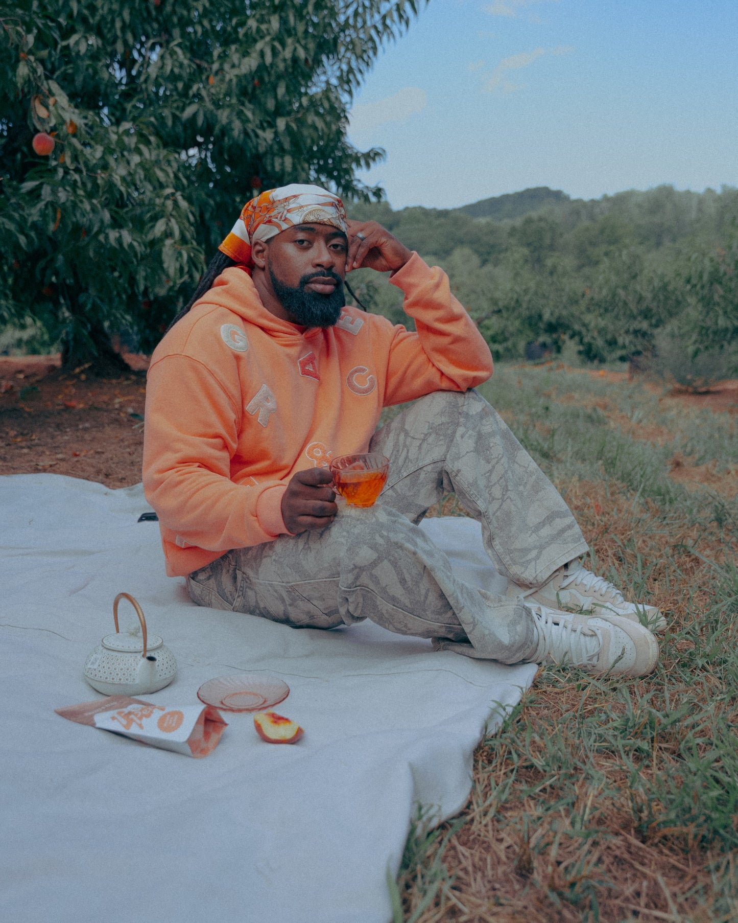 Man in an peach hoodie sitting on a blanket outdoors, Drinking tea