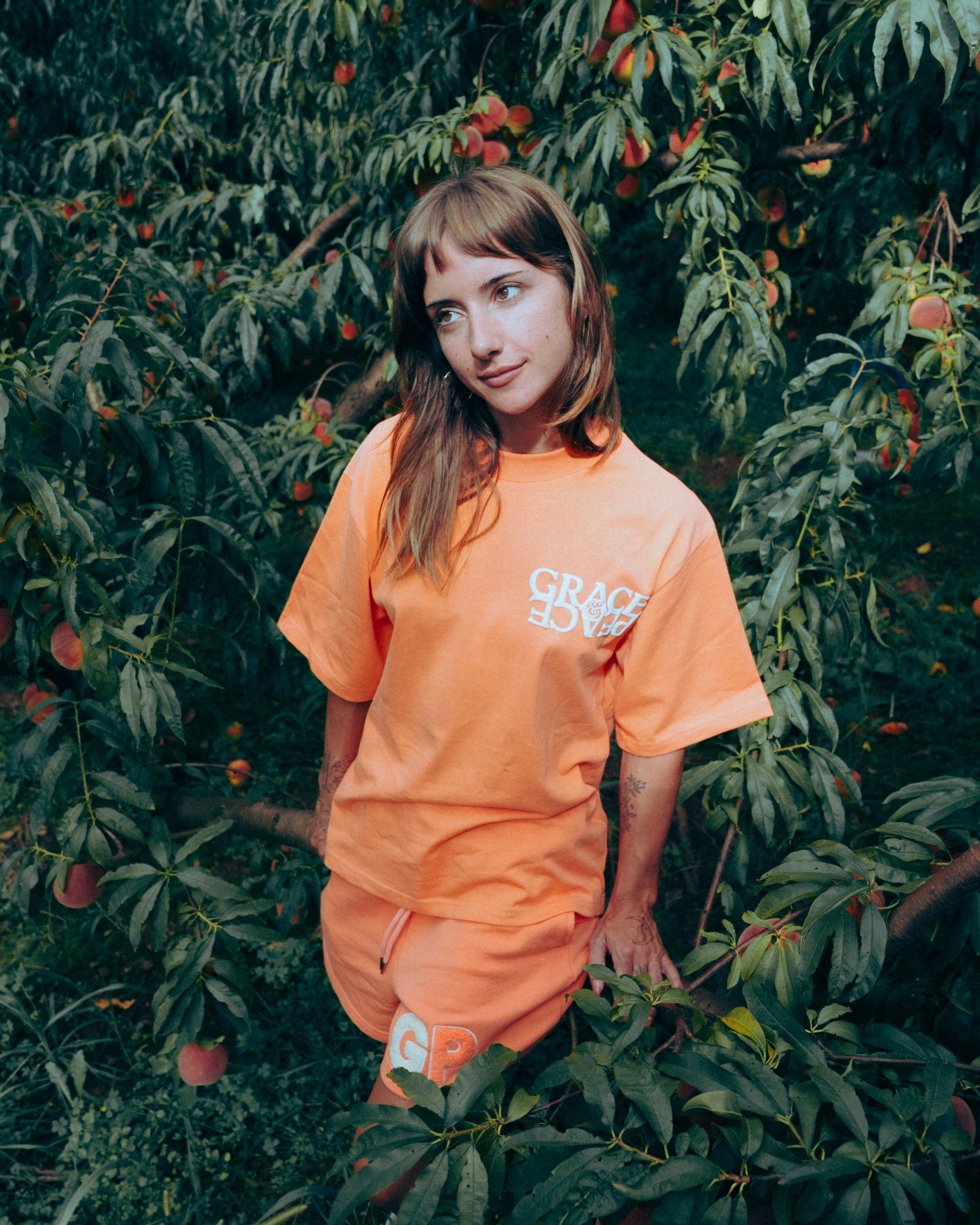 Person wearing an Peach outfit with 'Grace&peace' text, standing in a peach orahcard 